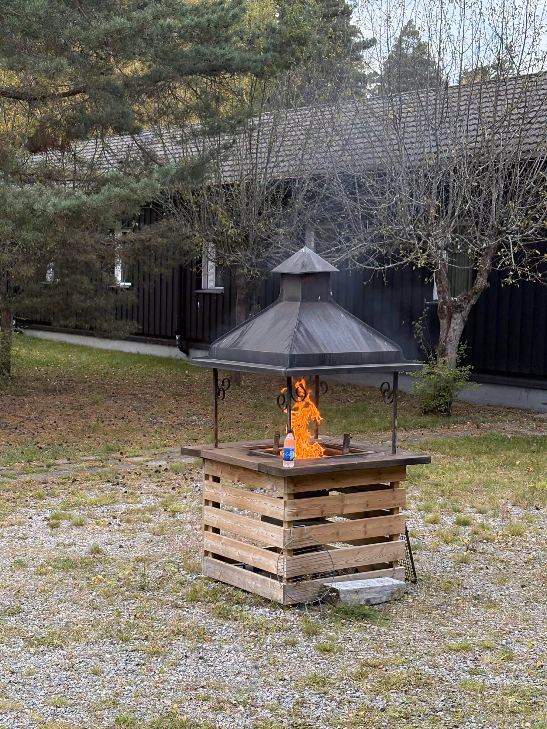 En brasa som brinner i en eldkorg med tak av metall, på gruset framför den svarta panelfasaden.