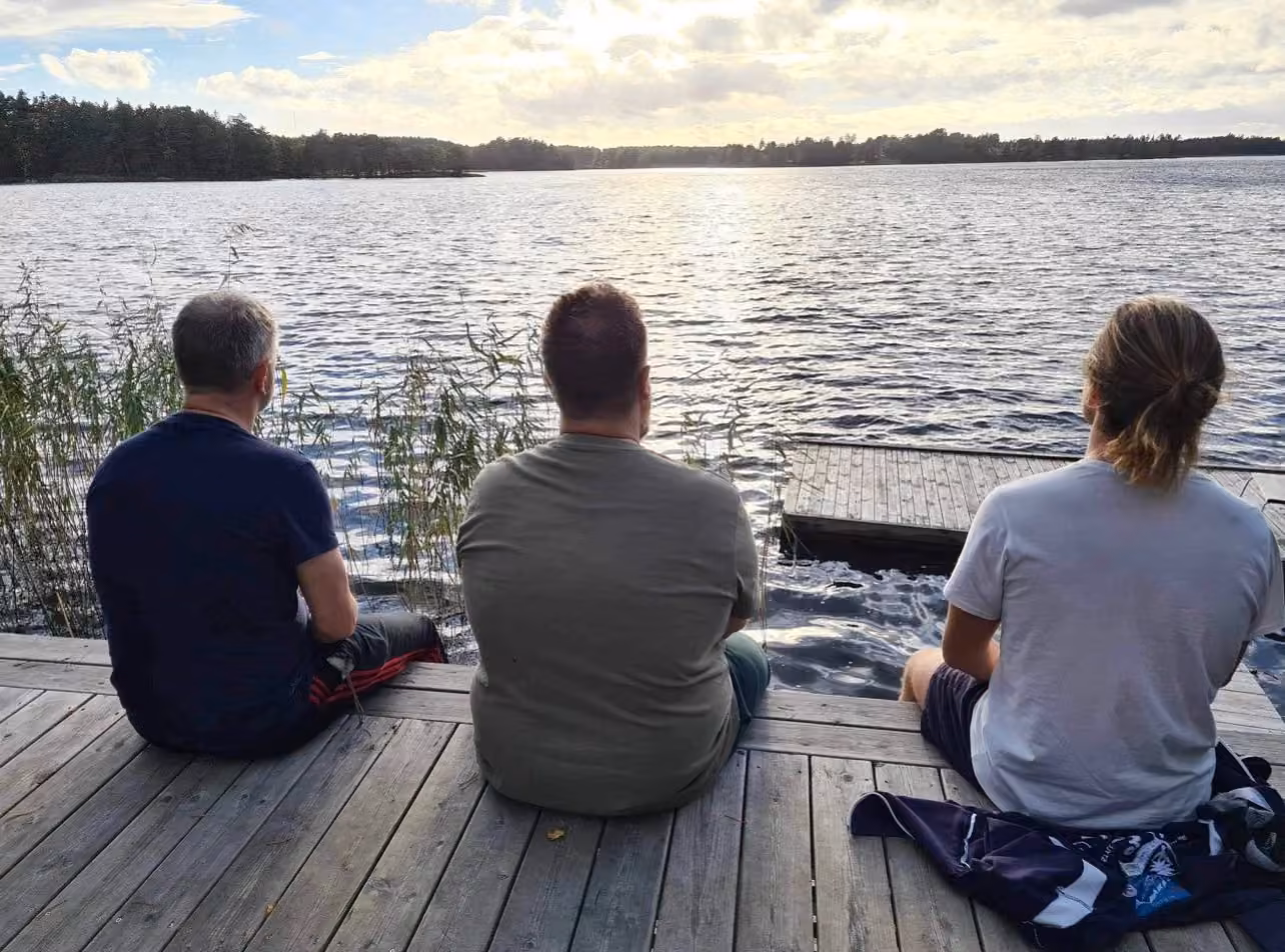 Tre personer sitter bredvid varandra på bryggan med ryggen mot kameran och ser ut över Båven, med vass på båda sidor och kvällssol över vattnet.