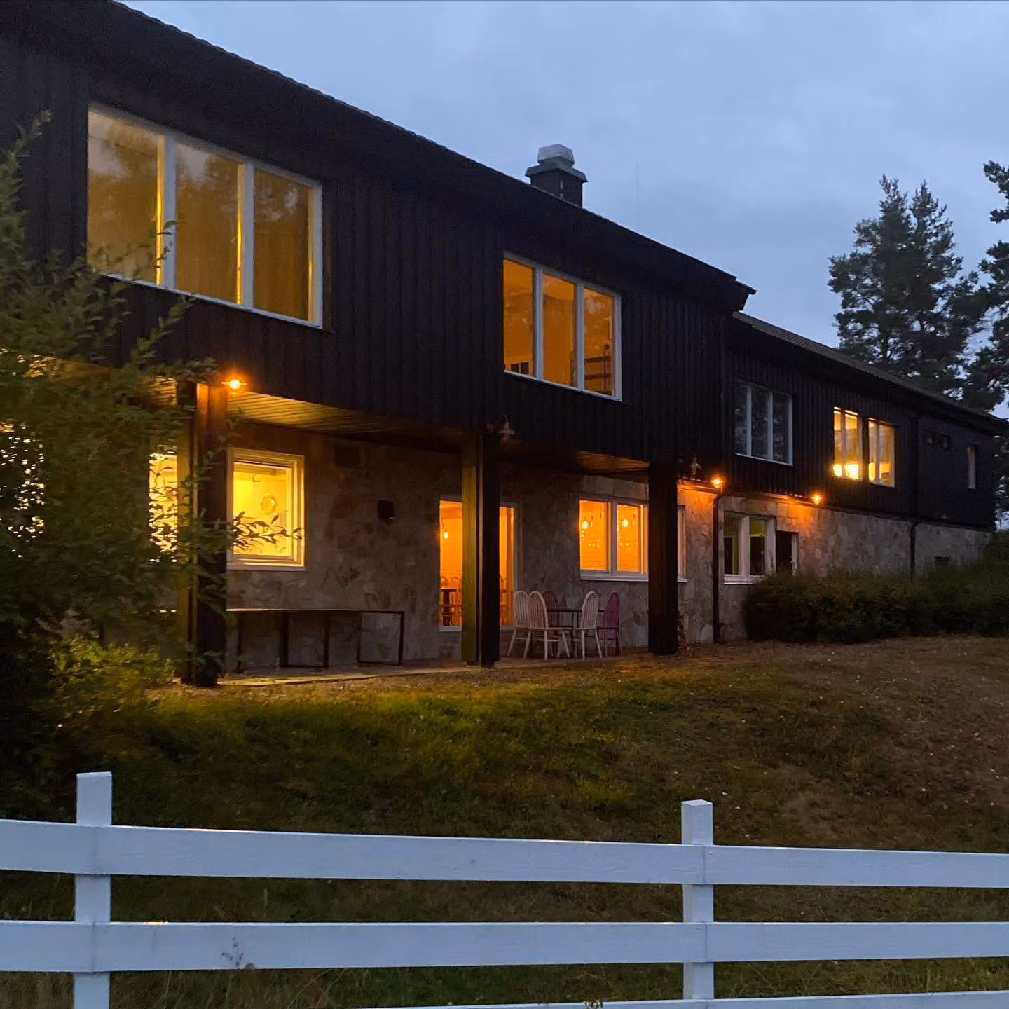 Kursgården i skymning. Fönsterraden lyser varmt gul mot den svarta träpanelen, ett vitt staket löper genom förgrunden och kvällshimlen är blå över taket.
