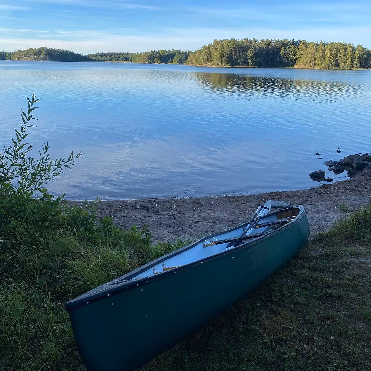 En grön kanot uppdragen på strandkanten, med spegelblank sjö och skogsklädd horisont bakom.