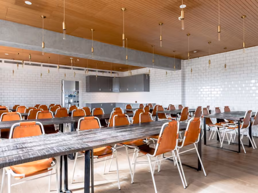 Matsal med långbord i grovt trä, orange läderstolar och panelat trätak med hängande mässingslampor.