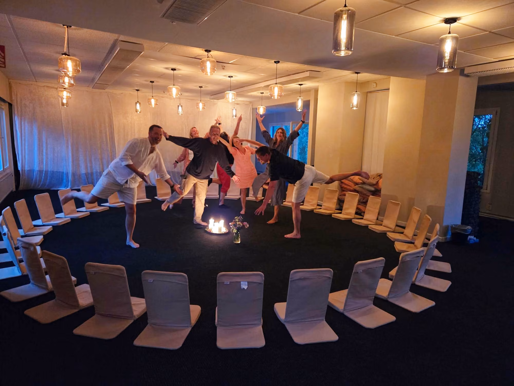 En grupp står på ett ben i en balansövning inne i ringen av meditationsstolar, i kvällsljus från glödlampor i taket och med tända ljus på golvet mitt i ringen.