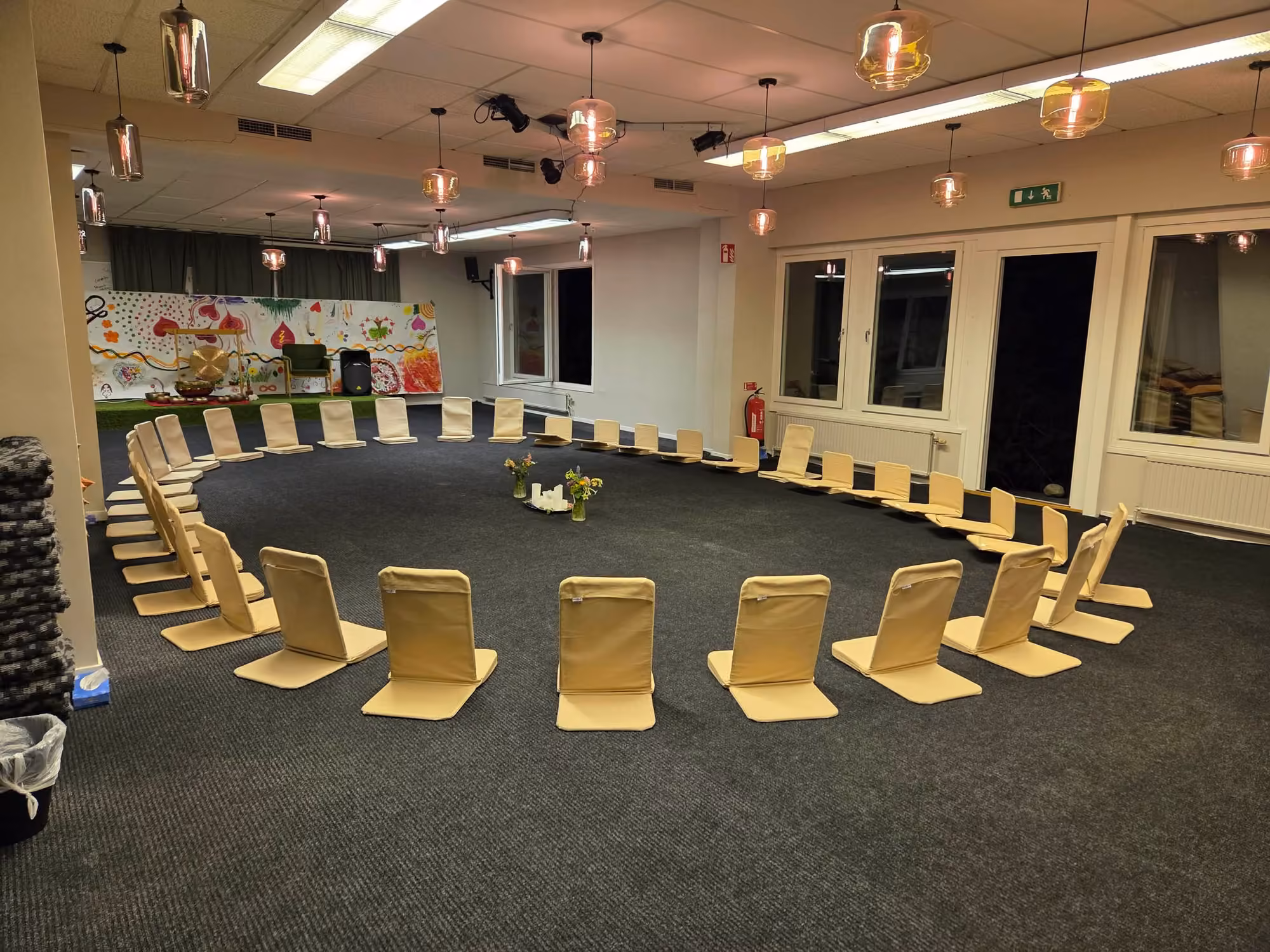 Salen på kvällen, med meditationsstolar i en vid ring, tända ljus och blommor mitt på golvet och varma glödlampor i taket.