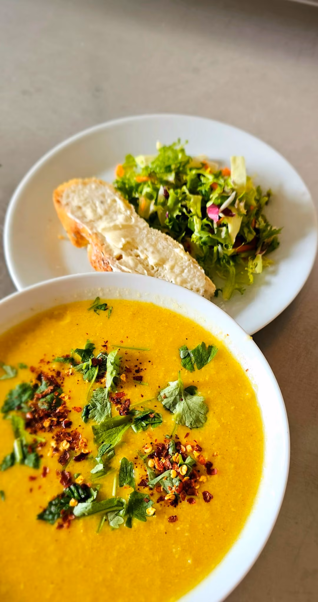 En djup tallrik gul rotfruktssoppa med koriander och chiliflingor, bredvid en tallrik med bröd och grönsallad.