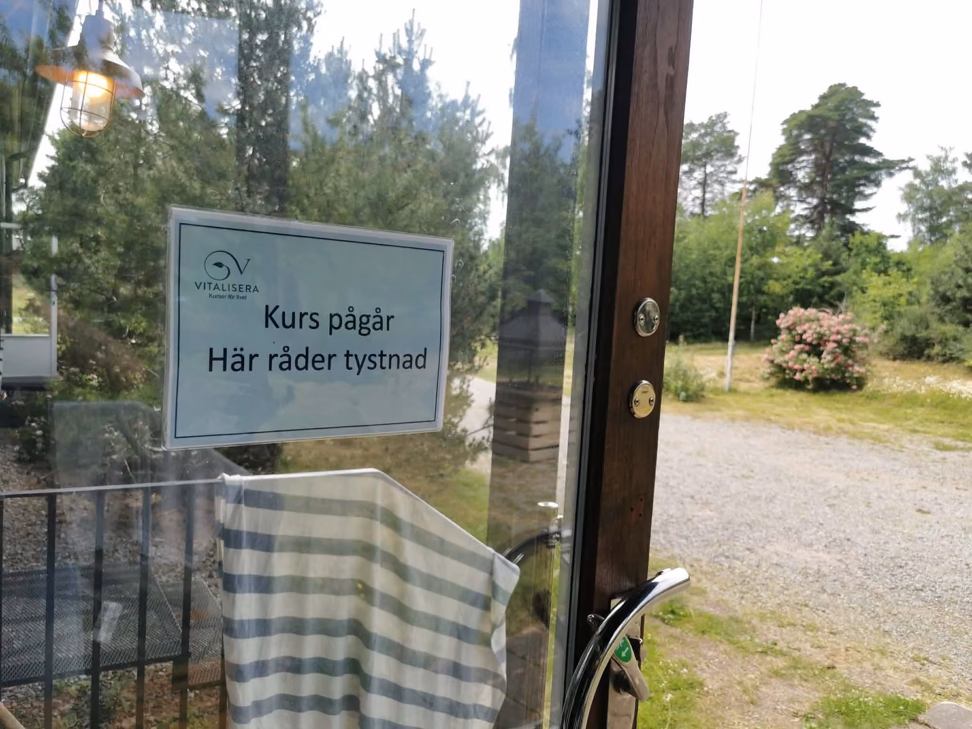 En laminerad skylt på insidan av en glasdörr med texten Kurs pågår, här råder tystnad. Genom glaset syns grusplanen, tallarna och en blommande buske.