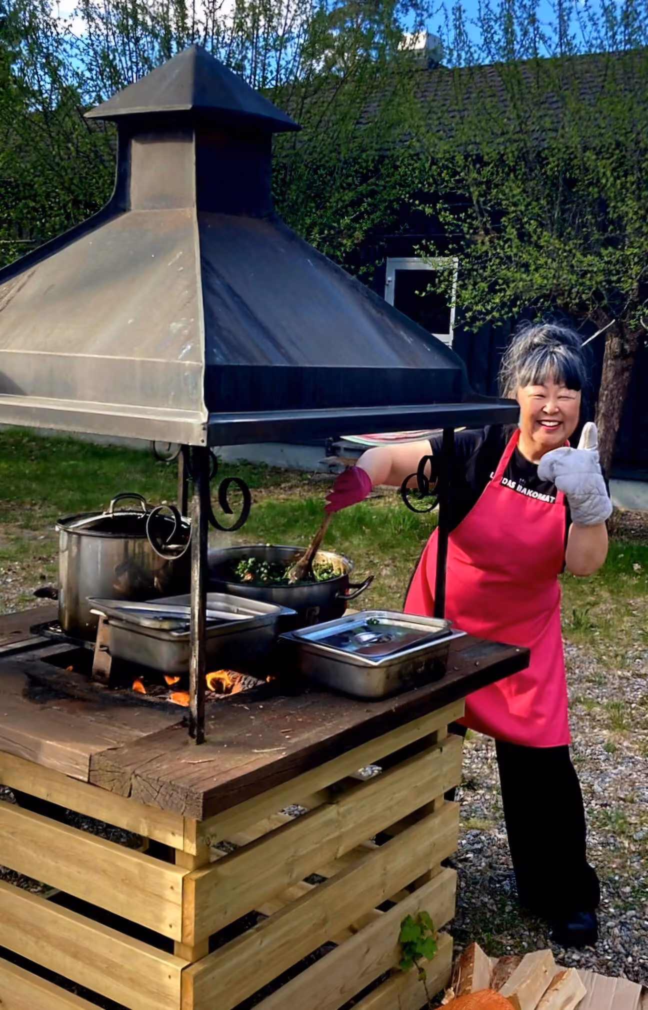 En kock står vid en gryta över öppen eld utomhus, med björkar i vårgrönt bakom.