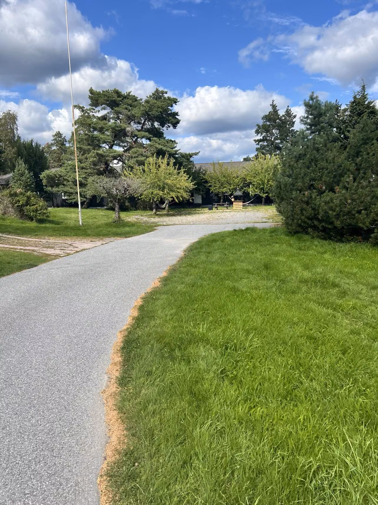 Vägen svänger in förbi husen med gräs på båda sidor, tallar och en flaggstång till vänster och skogen bakom.